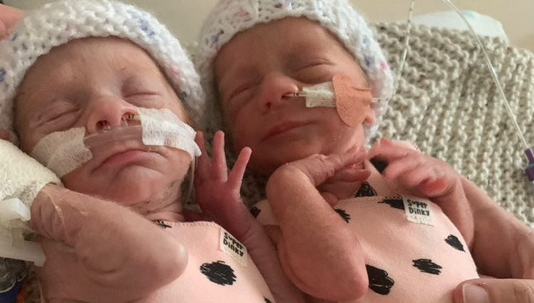 Two newborn babies in pink outfits with black spots, wearing hospital caps and receiving oxygen therapy.